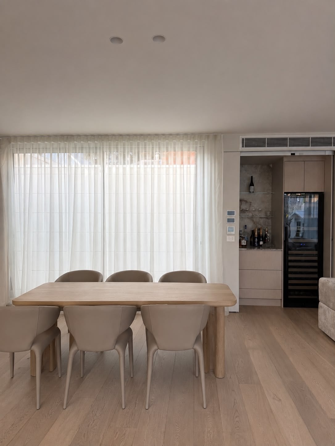 Dining room with a table and chairs before sheer curtains, a wine fridge set into the joinery, Port Melbourne